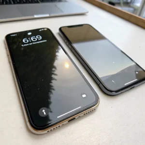 Two iPhones placed on a desk with a laptop in the background. One iPhone shows a lock screen reflecting a tree and sunset.