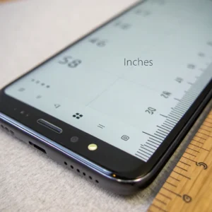 Smartphone screen displaying a measuring app in inches next to a physical ruler on a fabric surface.